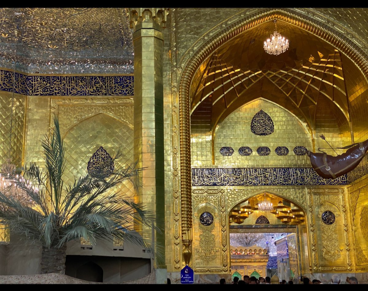 Interior of a grand mosque with ornate gold-tiled arches, decorative calligraphy, geometric patterns, palm plants, and a chandelier